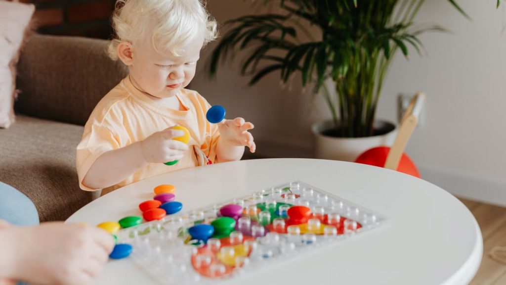 A toddler with white hair playing with colorful toys on a round table indoors, enjoying playtime.
